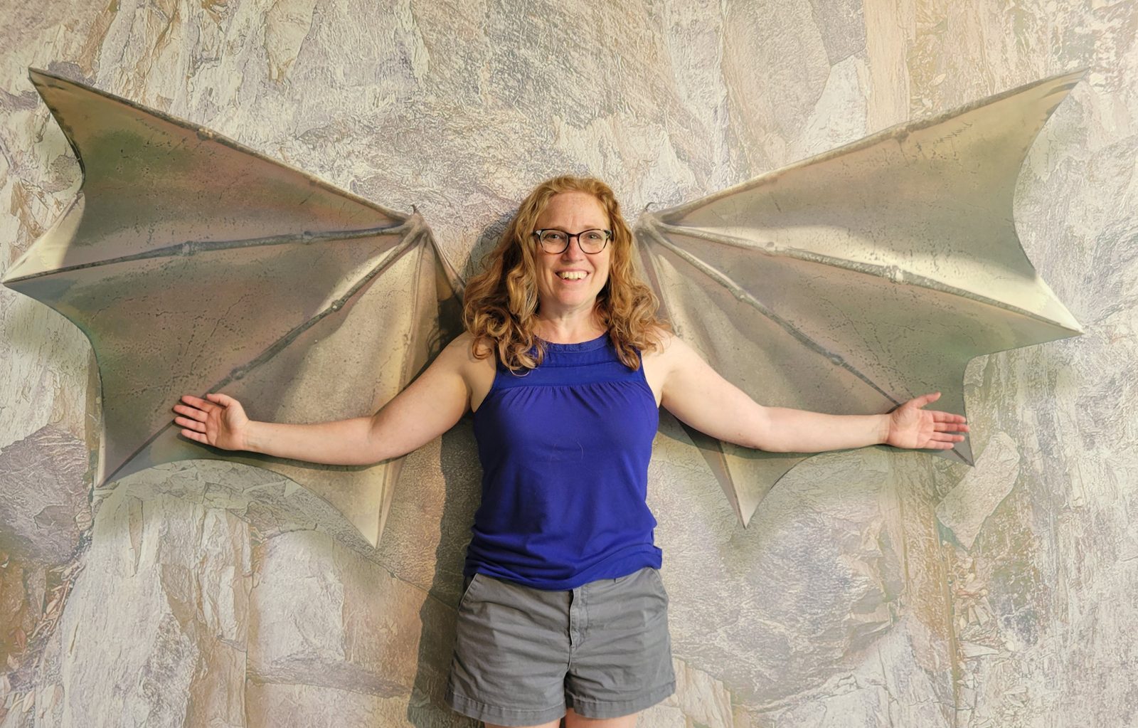 Stefani Pollack smiling in front of a wall mural of giant bat wings, arms spread wide